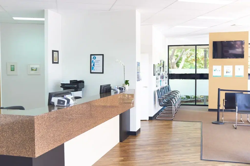 Modern medical office reception area in Redland Bay with a brown counter, chairs lined up along the wall, a printer, framed artwork, and large windows letting in natural light—an inviting space for Ear Relief consultations.